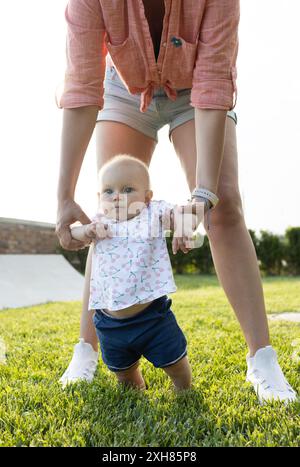 Ritratto di un bambino che fa i suoi primi passi sull'erba. madre felice che tiene le mani del suo bambino all'aperto nel parco. Stile di vita. La felicità Foto Stock