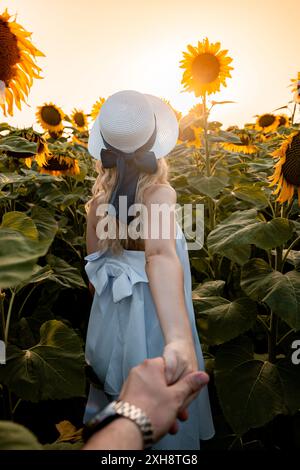 Una donna con un cappello bianco e un vestito azzurro cammina attraverso un campo di girasoli, tenendo la mano di qualcuno dietro di lei. Il sole che tramonta fa caldo Foto Stock