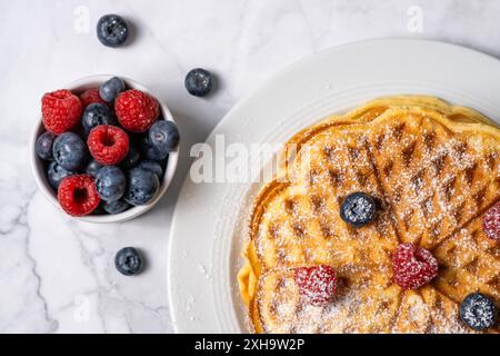 Vista dall'alto del waffle belga e mix di lamponi e mirtilli su sfondo in marmo. Deliziosa colazione estiva. Una pila di waffle rotondi fatti in casa Foto Stock