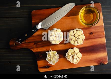 Preparazione dei bulbi d'aglio da tostare in olio d'oliva: Teste d'aglio fresche con il fondo tagliato su un tagliere di legno Foto Stock