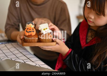 Colpo ritagliato di bambina che decora i cupcake di Halloween con mostri diversi Foto Stock