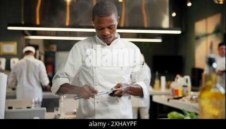 Uomo nero, chef e serio con affilacarichi in cucina al ristorante per cene raffinate, verdure e creatività. Dipendente, professionista e fornello Foto Stock