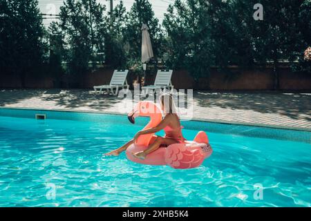 Una donna è seduta su un galleggiante rosa di fenicottero in una piscina. La scena è luminosa e allegra, con la donna che si gode il suo tempo in acqua. Foto Stock