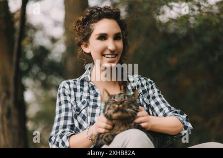 una giovane donna con i capelli ricci e una camicia a scacchi contiene un gatto tabby molto contento. Entrambi sembrano felici, con il gatto che sembra che stia godendo di una spa Foto Stock