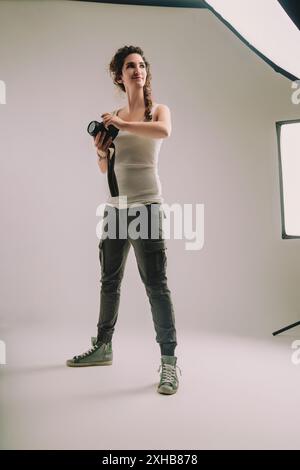 Una giovane donna con i capelli ricci intrecciati, vestita con una capote senza maniche e pantaloni cargo, tiene una macchina fotografica in uno studio. La sua posizione sicura e il suo rifiuto di espressione Foto Stock