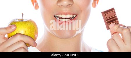 Bambino che tiene in mano una mela e un cioccolato. Bambino carino che sceglie tra mele e dolci. Cibo sano, denti buoni. Cibo cattivo cattivo Foto Stock