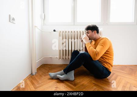 Un uomo con un maglione arancione siede sul pavimento vicino a un radiatore, bevendo da una tazza bianca. Indossa jeans blu e calzini grigi. Il pavimento è fatto di W. Foto Stock