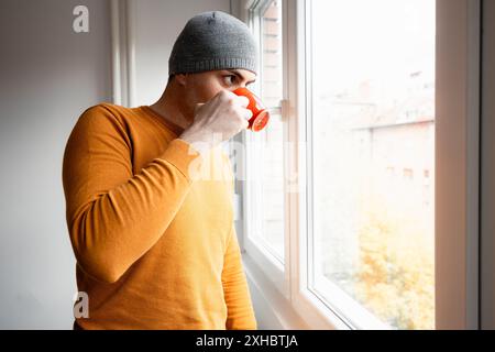 Un uomo con un berretto grigio e un maglione arancione si trova vicino a una finestra, tenendo una tazza rossa e guardando fuori. Foto Stock