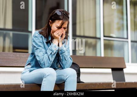 Una donna siede su una panchina fuori da un edificio, piangendo e tenendo la faccia. Indossa una camicia blu e un jeans. Foto Stock