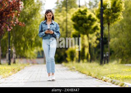 Una giovane donna che indossa una giacca in denim e un jeans cammina lungo un sentiero lastricato in un parco, guardando verso il basso il suo telefono. Foto Stock