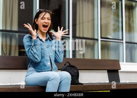 Una giovane donna con una giacca in denim si siede su una panchina, gridando di sorpresa con le mani alzate. Foto Stock