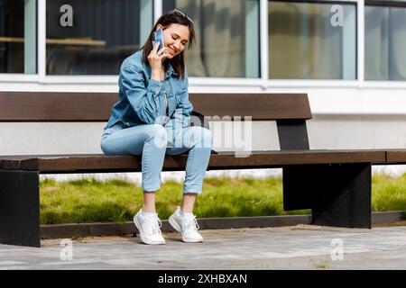 Una giovane donna che indossa una giacca in denim e un jeans blu siede su una panchina, sorride e parla al cellulare. Foto Stock