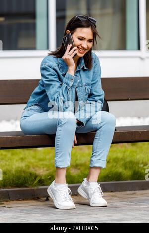 Una giovane donna che indossa una giacca in denim e un jeans siede su una panchina, sorride e parla al telefono. Foto Stock