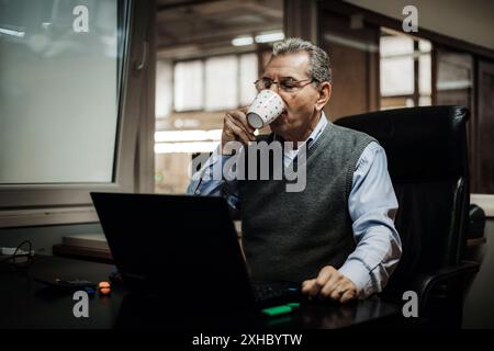 Un uomo anziano siede a una scrivania in ufficio, sorseggiando un caffè da una tazza bianca mentre utilizza un computer portatile. Foto Stock