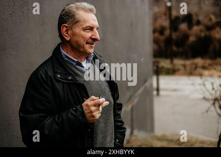 Un uomo con giacca nera e maglione grigio si appoggia contro una parete grigia, guardando lateralmente mentre fuma una sigaretta. Foto Stock