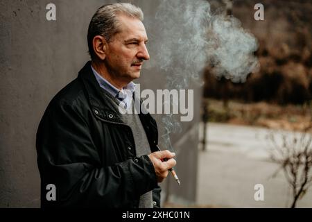 Un uomo anziano con una giacca nera e una camicia blu, sta fuori al freddo, espirando fumo da una sigaretta. Foto Stock