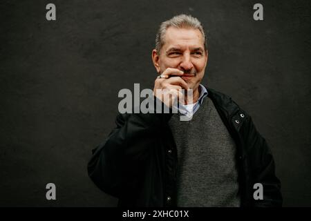 Un uomo con i capelli grigi e i baffi, con una giacca nera, sorride alla telecamera. E' in piedi di fronte a un muro oscuro. Foto Stock