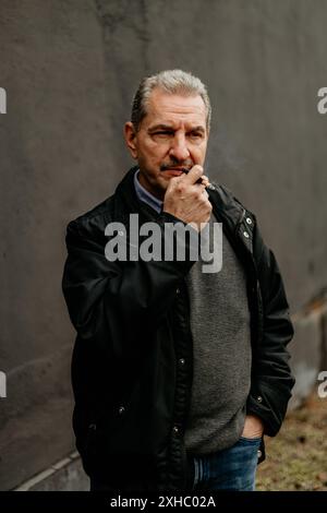Un uomo che indossa una giacca nera e un jeans blu si trova di fronte a una parete grigia. Ha i baffi e i capelli grigi. Sta tenendo una sigaretta in mano e.. Foto Stock