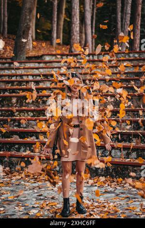Una donna con un cappotto marrone sorride mentre si trova in un parco, circondato da foglie autunnali cadenti. Foto Stock