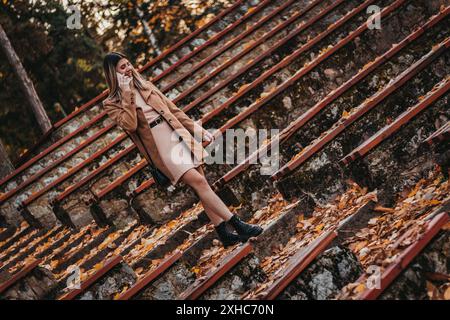 Una donna con un cappotto marrone sta su gradini di pietra, parla al telefono, con foglie cadute intorno a lei. Foto Stock