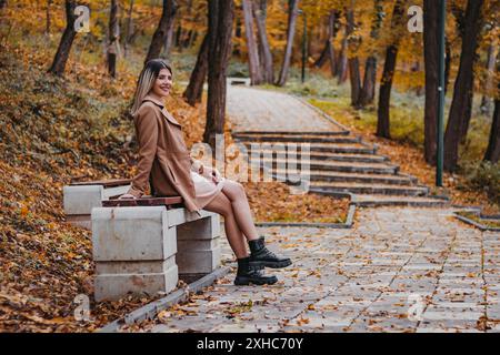 Una giovane donna che indossa una giacca marrone e un vestito rosa siede su una panchina in un parco. Sta guardando verso la telecamera. Il terreno è coperto in autunno Foto Stock