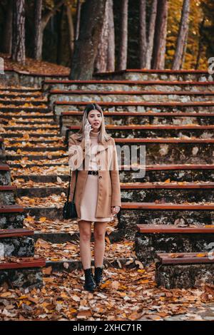 Una donna che indossa un cappotto marrone e un vestito rosa si erge su una serie di gradini ricoperti di foglie cadute, parlando al telefono. Foto Stock