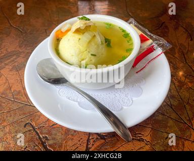 Una ciotola di zuppa di polpette di matzo si trova su un piatto bianco con un cucchiaio e una confezione di cracker saltini. La zuppa è guarnita con verdure tritate ed erbe aromatiche, Foto Stock