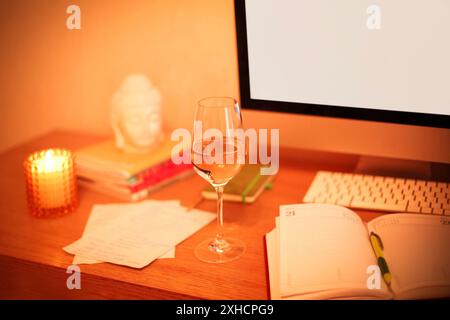 Bicchiere di vino collocato sul tavolo vicino al notebook e al monitor del computer con schermo vuoto la sera in ufficio Foto Stock