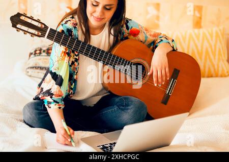 Crop musicista femminile con chitarra acustica e portatile che prende appunti mentre si siede a letto e scrivere canzone a casa Foto Stock