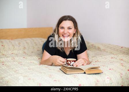 Donna di mezza età felicissima in abbigliamento domestico sdraiata su un comodo letto in camera da letto con una tazza di caffè e un libro interessante mentre si guarda la macchina fotografica Foto Stock