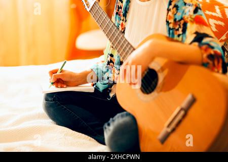 Crop musicista femminile con chitarra acustica e portatile che prende appunti mentre si siede a letto e scrivere canzone a casa Foto Stock