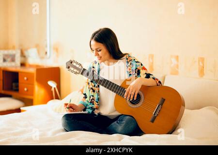 Crop musicista femminile con chitarra acustica e portatile che prende appunti mentre si siede a letto e scrivere canzone a casa Foto Stock