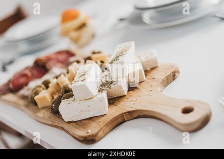 Un tagliere di legno siede su un tavolo bianco, riempito con una selezione di formaggi, olive e salumi. Foto Stock