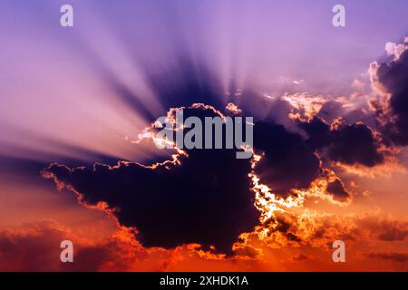 Un tramonto vibrante con raggi dorati di luce che penetrano attraverso una grande nuvola scura, illuminando le nuvole circostanti in un ardente bagliore arancione. Foto Stock