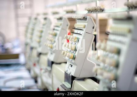 Diverse macchine da ricamo industriali sono allineate in una fabbrica, mostrando le bobine di filo colorato e gli aghi nella loro posizione di lavoro. Foto Stock