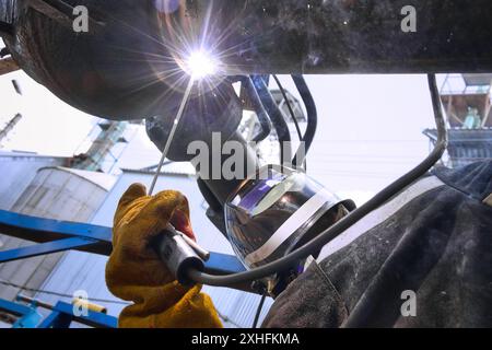 Una saldatrice che indossa una maschera protettiva e guanti collega due tubi utilizzando una torcia per saldatura. La luce brillante dell'arco di saldatura illumina l'area di lavoro e la mano con i guanti della saldatrice che tiene la torcia di saldatura. Foto Stock