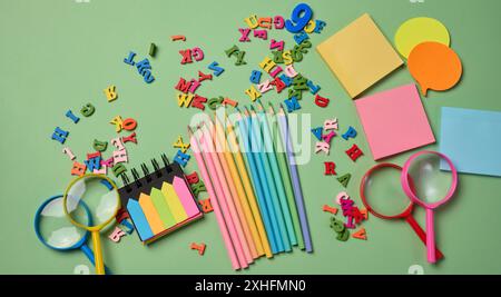 Cancelleria disposta su sfondo verde. Matite multicolore , appiccicose e lettere di legno, preparazione allo studio o al lavoro, organizzazione dello spazio di lavoro Foto Stock