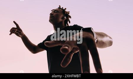 Gen Z uomo con dreadlock balla energicamente in uno studio. Occhi chiusi, si muove al ritmo della musica, le mani fluiscono. Indossa una camicia rosa, lui Foto Stock