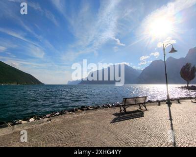 Nago-Torbole, Lago di Garda, Italia - 13 luglio 2024: vista del pittoresco Lago di Garda a Torbole, in Italia, con una panchina solitaria sulla riva, circondato da imponenti montagne e dalla luce solare brillante *** Blick auf den malerischen Gardasee a Torbole, Italien, mit einer einsamen Bank am Ufer, umgeben von beeindruckenden Bergen und strahlendem Sonnenlicht Foto Stock