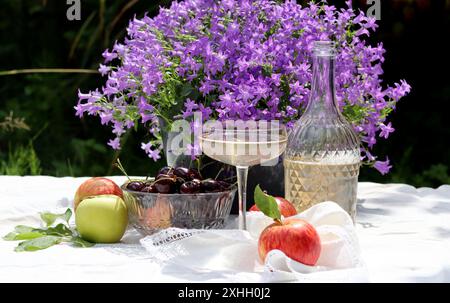 Tavolo da picnic con vino, mele, ciliegie e fiori nel giardino estivo. Bella composizione di frutta e fiori di stagione. Foto Stock