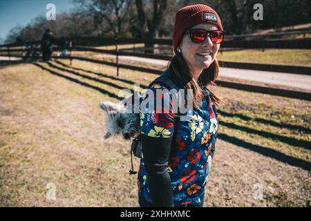 Una donna con un abbigliamento colorato e un berretto rosso che cammina all'aperto con un piccolo cane nel suo zaino in una giornata di sole Foto Stock