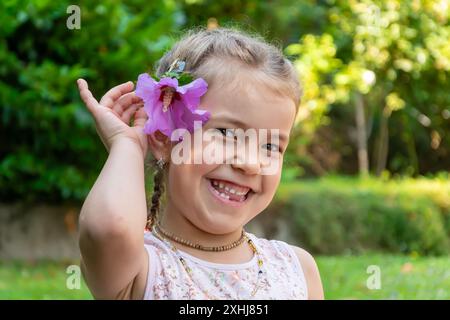 Ritratto di una bambina di 6 anni che ride con un fiore tra i capelli sullo sfondo di un giardino fiorito. I denti del latte della ragazza sono caduti. Foto Stock