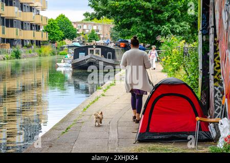 Una signora cammina il suo cane davanti a una tenda posta sotto un ponte sul percorso lungo il braccio di Paddington del Grand Union Canal a Londra, Regno Unito Foto Stock