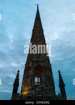 L'alta guglia della chiesa è alta contro un cielo nuvoloso, illuminato dalla calda luce del sole che tramonta Foto Stock