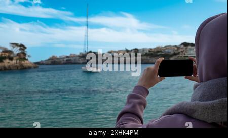 Una donna usa il suo smartphone per fotografare la splendida costa mediterranea mentre una barca a vela è ancorata nelle vicinanze Foto Stock