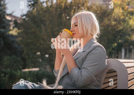 Una donna bionda siede su una panchina bevendo caffè da una tazza gialla. Indossa una giacca grigia e ha i capelli in una coda di cavallo. La scena è tranquilla Foto Stock