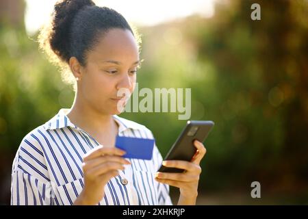 Donna nera seria che acquista online usando il telefono e la carta di credito in un parco Foto Stock