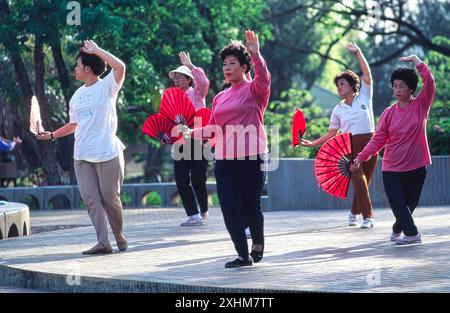 Donne thailandesi di mezza età che praticano Tai-chi all'aperto nel Lumpini Park, Bangkok, Thailandia, tenendo in mano i tradizionali fan di carta, indossando top colorati e pantaloni neri, catturate con aggraziato movimento durante una sessione mattutina di sole. Foto Stock
