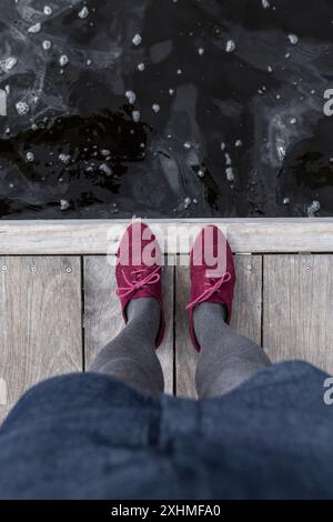 I piedi di una persona indossati con scarpe rosse e collant grigi in piedi su un molo di legno Foto Stock