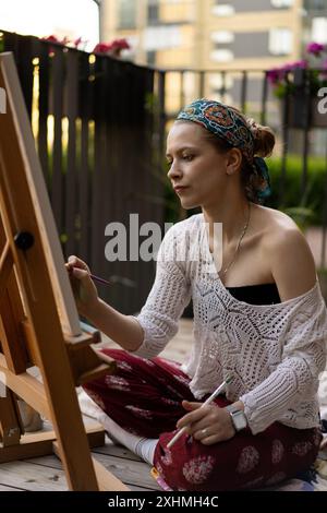 Artista donna dipinge su tela, pitture a olio. Cavalletto in terrazza, in estate. Foto Stock